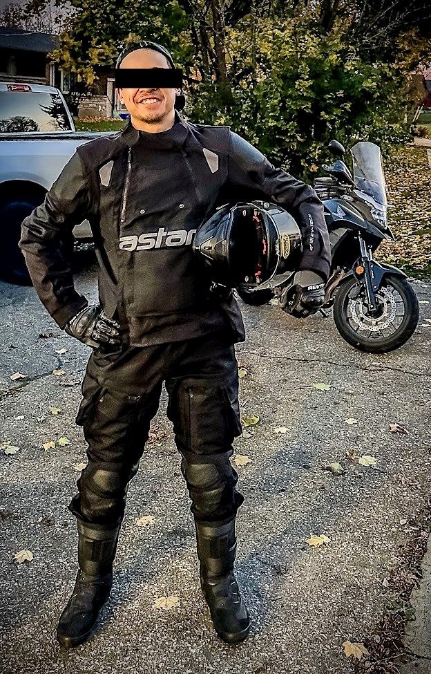 Alex in full riding gear holding a helmet beside his motorcycle